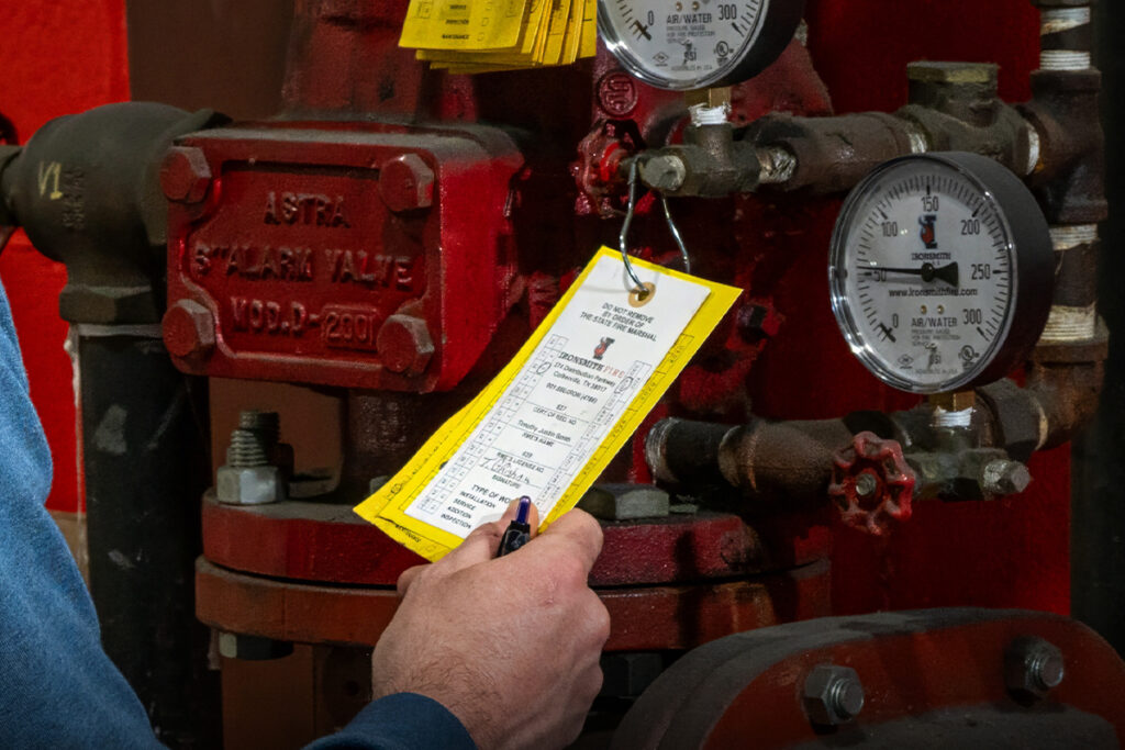 Technician filling out an inspection tag on a red fire sprinkler alarm valve during fire protection ITM