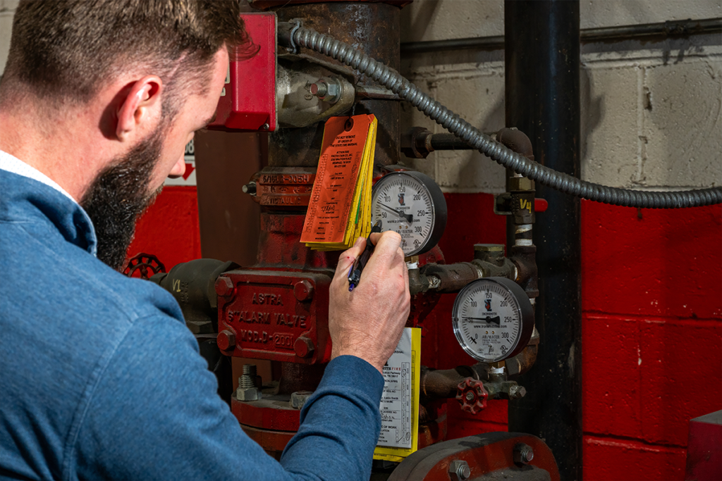 Technician writing on an inspection tag at a fire sprinkler riser alarm valve